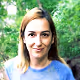 Woman with light brown hair wearing a blue shirt, smiling outdoors with greenery in the background.