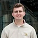 Young man with short brown hair wearing a light beige polo shirt, smiling in an outdoor urban setting.