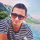 Man wearing sunglasses and a black and white striped shirt with a mountainous landscape and body of water in the background.