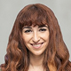 Smiling woman with long curly reddish-brown hair and bangs, wearing a white top.
